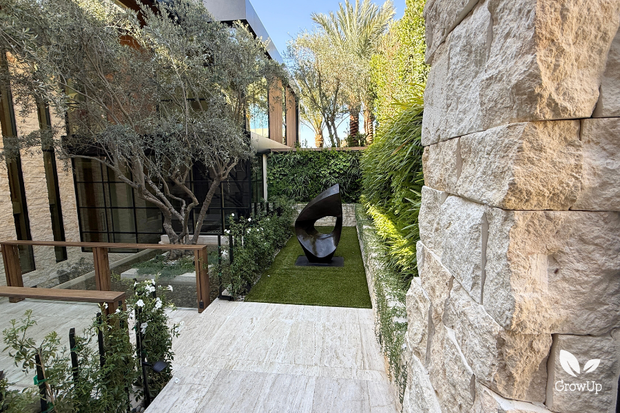 An oblique view of a modern luxury courtyard garden, framed on the right by a large, textured limestone block wall. A light travertine pathway leads past a patch of artificial turf featuring a large, dark geometric sculpture. In the background is a multi-story modern home with extensive glass windows and a large, mature olive tree. A high hedge provides a backdrop, with a section of living green wall visible near the sculpture.