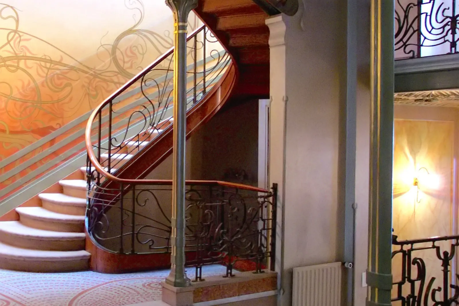 An Art Nouveau residential interior featuring a sweeping wooden spiral staircase with decorative wrought iron railings shaped like organic vines, alongside muted floral wall frescoes.