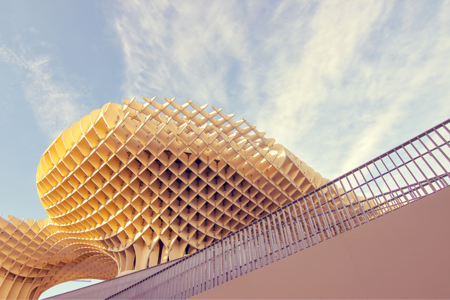 A low-angle view of the Metropol Parasol timber structure in Seville, featuring a massive, flowing wooden lattice canopy shaped like giant interconnected mushrooms against a blue sky.