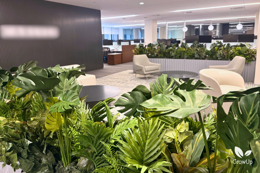 A spacious, modern office lounge and breakout area with comfortable grey and white armchairs, low circular tables, and dense corporate planter boxes filled with lush tropical greenery and Monstera deliciosa leaves.