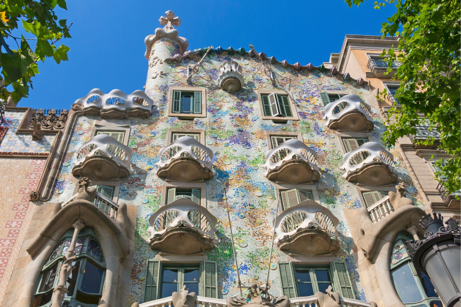 A full view looking up at the colorful mosaic facade of Antoni Gaudi's Casa Batllo in Barcelona under a blue sky, displaying organic skull shaped balconies and a wave shaped roofline.