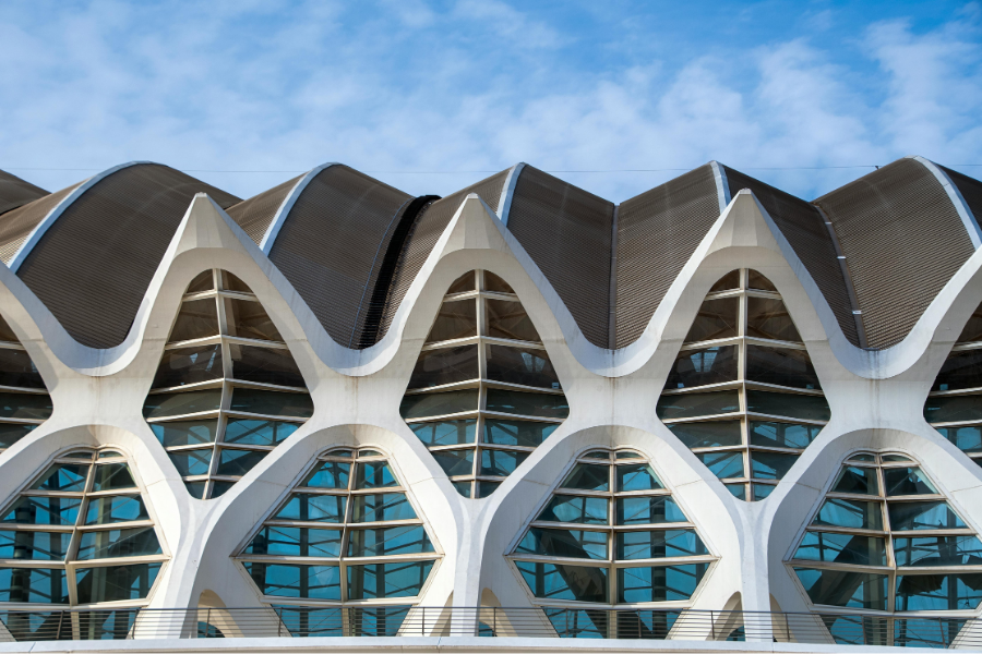 A low-angle shot of a modern white building facade featuring repetitive, skeletal, pointed arches and geometric glass windows that mimic a biological spine or rib cage.
