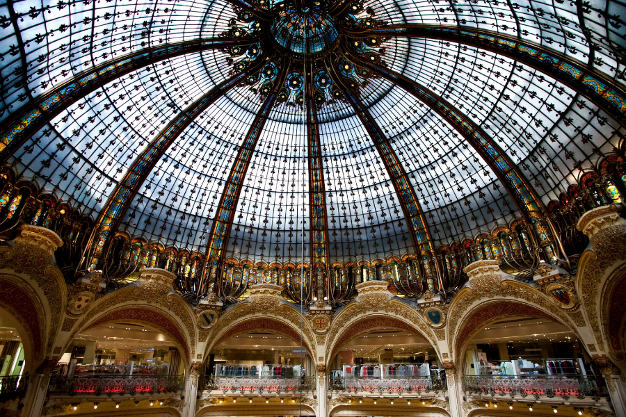 A massive, intricate Art Nouveau stained glass and iron dome ceiling flooding a grand multi level building interior with natural light.