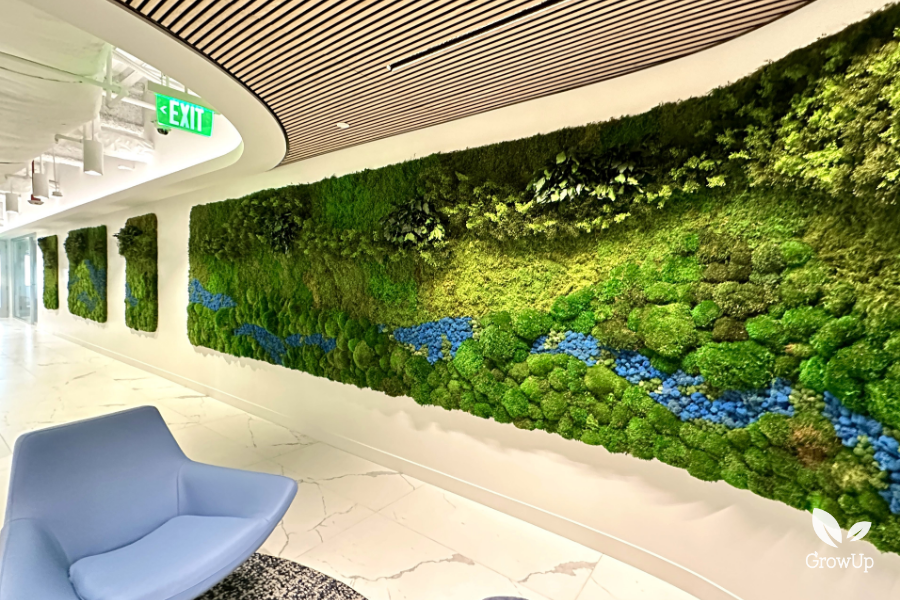 A long office corridor featuring a massive, textured green moss wall installation inlaid with a winding stream pattern of bright blue moss.