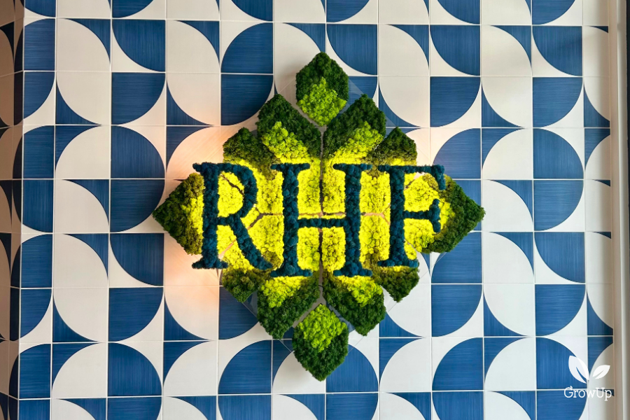 A diamond-shaped company logo made of green and yellow moss with blue moss lettering spelling out "RHF," mounted on a blue and white geometric tiled wall.