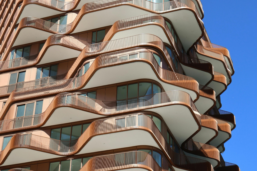 A close-up exterior view of a modern high-rise building featuring fluid, undulating bronze balconies that curve irregularly on each floor to look like layered rock formations.