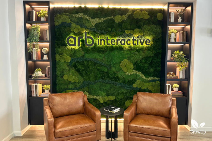 A welcoming office reception area featuring two brown leather armchairs, a small black side table with books, and a large biophilic preserved moss wall in the background with the backlit logo "arb interactive" and flanking black shelving units with various decorative items.