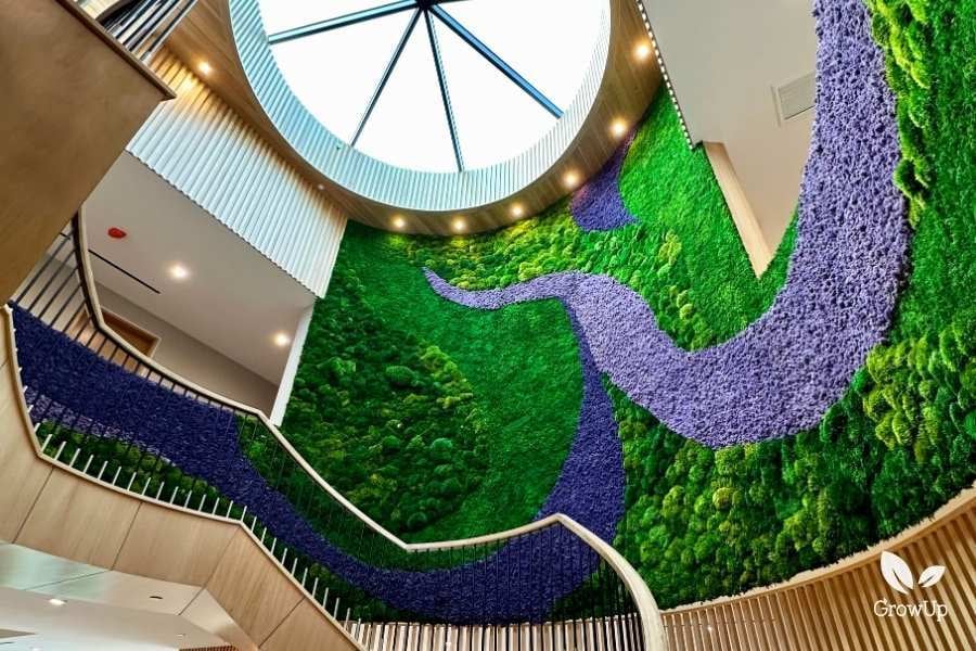 A multi-story atrium staircase featuring a massive, curved green moss wall with a flowing, river-like pattern made of purple moss stretching toward a circular skylight.